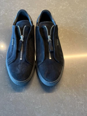 Oanvända Axel Arigato Clean 90 Zip svart mocka - Stilrena svarta sneakers från Axel Arigato i mocka med coola silverfärgade dragkedjor istället för snörning. Låg modell med diskret logga på sidan. Perfekta för dig som gillar minimalistisk och modern design.