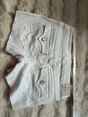Vita jeansshorts från True Religion - Snygga vita jeansshorts från True Religion med klassiska bakfickor och silverfärgade knappar. Shortsen har bälteshällor och femficksdesign. Perfekta för varma dagar och ger en fräsch look till din outfit.