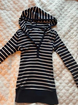 Randig hoodie från Hollister marinblå - Snygg marinblå och vitrandig hoodie från Hollister med knappar vid halsen och lång ärm. Klassisk logga på bröstet och stor huva. Mjuk och stretchig bomullsblandning som passar perfekt till jeans eller leggings.