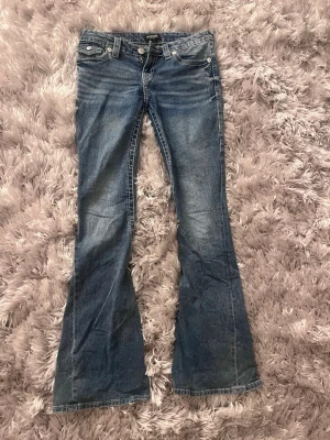 Blå bootcut jeans från Replay - Snygga blå bootcut jeans från Replay med slitna detaljer och markerade sömmar. Jeansen har klassisk femficksdesign, knappar på bakfickorna och fransade inslag längs benen. Perfekta för en avslappnad och trendig look.