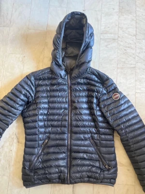 Svart dunjacka från Colmar - Snygg svart dunjacka från Colmar med huva och dragkedja framtill. Jackan har två fickor med dragkedja och ett Colmar-märke på ärmen. Lätt och smidig modell som håller värmen under kalla dagar.