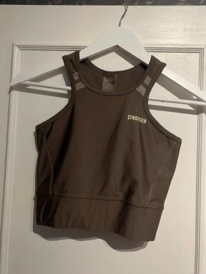Brun sport-bh från Stronger - Snygg brun sport-bh från Stronger med hög halsringning och brottarrygg. Tillverkad i stretchigt funktionsmaterial, perfekt för träning. Diskreta vita logotyper fram och bak samt små meshdetaljer vid axlarna för extra ventilation. I storlek xs❣️
