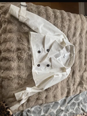 Vit cropped kappa med bälte - Trendig vit cropped kappa med dubbelknäppning och stora svarta knappar. Jackan har krage och bälte vid ärmsluten men ena armen har inte det för den har gått av men det går att knyta ändå. Vid magen finns det lite fläckar men det går säkert att tvätta, jag har provat den en gång men den var för stor, jag köpte den av en vän och hon har använt den några gånger, den köptes för 400kr men säljer för 150. 