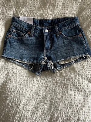 Blå jeansshorts från Weekday - Säljer ett par blå jeansshorts från Weekday, modell Venus Hotpants som är lowwastied. Shortsen har råa kanter, klassiska bakfickor och bälteshällor. Perfekta för varma dagar och varje tillfälle. Dem är ALDRIG använda. Dem perfekta sommarshortsen har ni här!