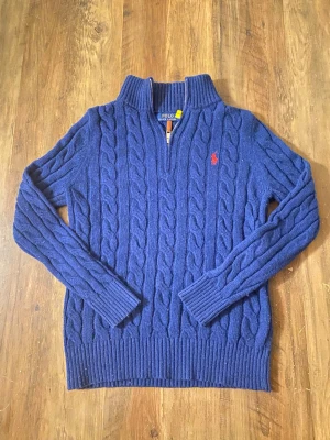 Blå kabelstickad tröja från Polo Ralph Lauren - Mörkblå kabelstickad tröja med halv dragkedja och hög krage från Polo Ralph Lauren. Tröjan har ett rött broderat logomärke på bröstet och är gjord i ett mjukt, värmande material. Perfekt för lager-på-lager och snygg till jeans.
