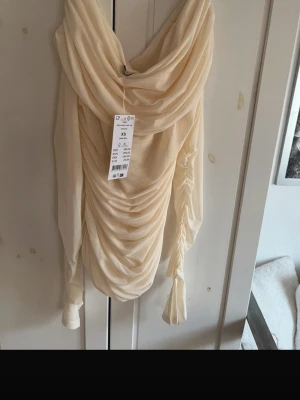 Ljus omlottklänning med drapering - Trendig ljus klänning från NA-KD med snygg drapering och omlott-detalj. Klänningen har långa ärmar och en elegant, rynkad design framtill. Perfekt för dig som vill sticka ut med en modern och stilren look.