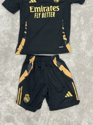 Real Madrid svart träningsset Adidas - Snyggt svart träningsset från Adidas med Real Madrid-logga och guldiga detaljer. Tröjan har korta ärmar, guldiga ränder på axlarna och tryck med 'Emirates Fly Better'. Matchande shorts med guldiga detaljer och klubbmärke. Perfekt för fotbollsträning.