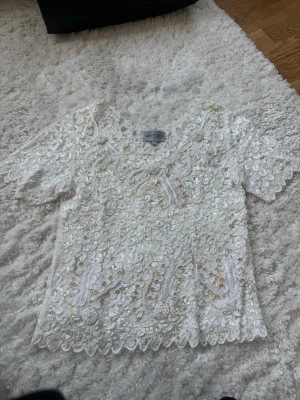 Vit paljettblus med spetsdetaljer - Supersöt vit blus från ART CIRCLE, täckt med glittriga paljetter och broderade spetsdetaljer. Kortärmad modell med v-ringning och scallop-kant. Perfekt för dig som vill sticka ut lite extra med en unik topp!