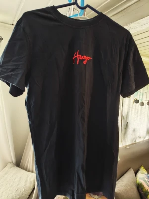 Svart t-shirt från Hugo med röd logga/M - Svart t-shirt från Hugo med röd broderad logga på bröstet. Klassisk rund halsringning och korta ärmar. Tillverkad i mjuk bomull som känns skön mot huden. Perfekt för en clean och stilren look.