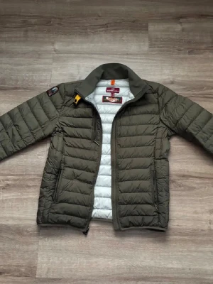 Olivgrön pufferjacka från Parajumpers - Snygg olivgrön pufferjacka från Parajumpers med quiltad design och grå insida. Jackan har hög krage, dragkedja framtill, två sidofickor med dragkedja och en gul detalj vid halsen. Perfekt för kalla dagar och riktigt trendig stil. Jackan har ett litet litet hål på armbågen som ej syns