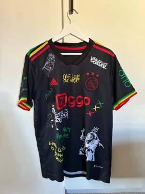 Svart Ajax x Bob Marley t-shirt Adidas - Unik svart t-shirt från Adidas i samarbete med Ajax och Bob Marley. Färgglada detaljer i rött, gult och grönt på ärmarna, tryck med Bob Marley, texter och Ajax-logga. Perfekt för dig som gillar fotboll och reggae. Storlek L ” men mindre i storlek” passar mig och jag är 181cm och väger ca 63kg den är lite oversized bara.