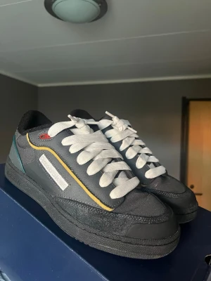 Reebok Club C Bulc - Snygga Reebok Club C 85 bulc sneakers i mörkgrått med vita snören, gula och blå detaljer samt svart sula. Skorna har klassisk låg siluett och är tillverkade i en mix av mocka och textil. Storlek 43 ganska true to size annars något större.  Väldigt bra skick endast sulan är lite dammig. Låda samt paketeringspapper kommer med