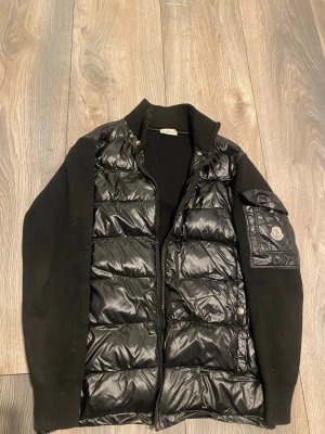 Svart dunväst från Moncler - Snygg svart dunväst från Moncler med quiltad framsida och ärmar i stickat material. Västen har dragkedja, hög krage och en cool ficka med Moncler-logga på ärmen. Perfekt för lager-på-lager och streetstyle.