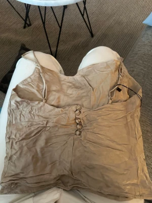 Beige linnetopp med knappar - Snygg beige linnetopp i satinliknande material med smala justerbara axelband och dekorativa knappar framtill. Perfekt att styla med jeans eller kjol för en trendig look.