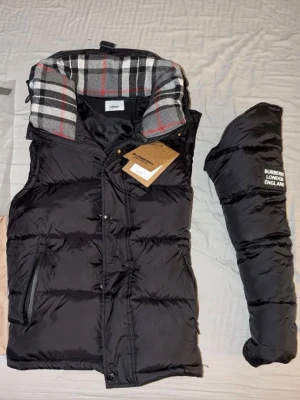 Svart dunväst från Burberry - Stilren svart dunväst från Burberry med klassiskt rutigt foder i grått, vitt och rött. Västen har dragkedja, knappar och två fickor framtill. Märkets logga syns på tillhörande fodral. Perfekt för lager-på-lager och snygg streetstil.