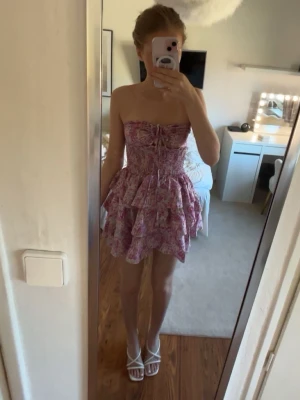 Rosa blommig sommar klänning  - Säljer nu denna fantastiska rosablommiga miniklänning. Modellen är axelbandslös med ett stretchigt material med fina volanger på kjolen som ger en jättesöt siluett. Helt perfekt för sommarens middagar på stranden eller semesterresor! 