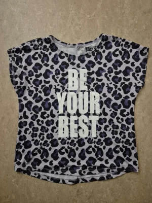 T-shirt - Cool t-shirt från Active med leopardmönster i svart och lila på grå botten. Stort vitt tryck framtill med texten 'BE YOUR BEST'. Kortärmad och i mjukt material, perfekt för en trendig vardagslook.