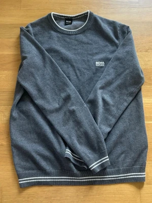 Grå stickad tröja från Hugo Boss - Mörkgrå stickad tröja från Hugo Boss med rund halsringning och vit logotyp broderad på bröstet. Tröjan har ribbstickade muddar och nederkant med vita ränder för en sportig touch. Perfekt för dig som gillar stilren design.