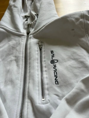 Vit hoodie från Sail Racing - Vit hoodie från Sail Racing med dragkedja och en extra bröstficka med dragkedja. Broderad logga på bröstet. Mjuk bomullskänsla och klassisk huva. Perfekt för en sportig och avslappnad stil. Lite smutsig 