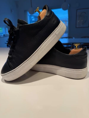 Nubikk Sneakers i premiumläder – stilrena & bekväma - Säljer ett par eleganta svarta sneakers från exklusiva nederländska märket Nubikk i storlek 41. Perfekta för dig som vill ha en stilren sneaker med hög komfort och premiumkänsla.  ✔ Tillverkade i kornigt premiumläder med snygg struktur✔ Minimalistisk design med rena linjer✔ Vit kontrasterande gummisula för en modern och tidlös look✔ Diskret Nubikk-logotyp på plösen✔ Bekväma och lätta att bära hela dagen  Färg: Svart med vit sulaStorlek: 41  Skoblock ingår ej i försäljningen. 