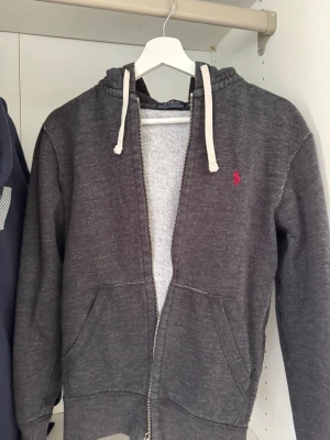 Mörkgrå hoodie från Polo Ralph Lauren - Mörkgrå hoodie med dragkedja från Polo Ralph Lauren. Klassisk design med vit dragsko i huvan och röd broderad logga på bröstet. Mjuk bomullsblandning och två fickor framtill. Perfekt för en avslappnad och stilren look.