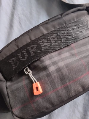 Svart rutig Burberry handväska - Snygg svart handväska från Burberry med klassiskt rutmönster i grått och röda detaljer. Väskan har en bred axelrem med Burberry-logga och dragkedja med orange zip-pull. Perfekt för dig som gillar streetstyle och vill ha något trendigt.