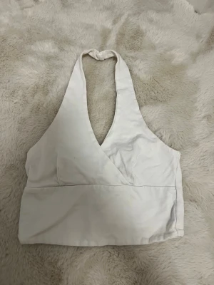 Vit omlott halterneck linnetopp - Snygg vit halterneck linnetopp med omlott-skärning och djup v-ringning. Toppen knyts i nacken och har en croppad passform som passar perfekt till höga jeans eller kjol. Enkel och stilren design som är lätt att matcha.