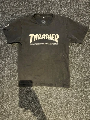Svart Thrasher t-shirt i bomull - Cool svart t-shirt från Thrasher med klassisk vit logga och texten 'Skateboard Magazine' på bröstet. Tillverkad i 100% bomull och har en avslappnad passform. Perfekt för dig som gillar streetwear och skatekultur.