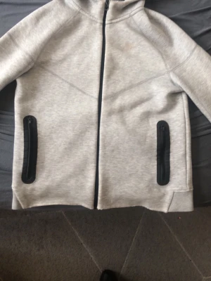 Grå Nike hoodie med dragkedja - Snygg grå hoodie från Nike med hel dragkedja framtill och svarta dragkedjeförsedda fickor. Klassisk Nike-logga på bröstet och huva för extra stilpoäng. Perfekt för en sportig och avslappnad look. Nike-loggan är även lite bortsliten på grund av att den har tvättats många gånger. Även på armbågarna så är det lite skrapmärken, men det är inget som man lägger märke till.