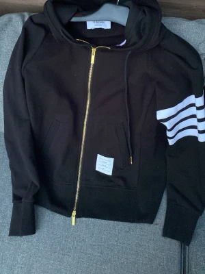 Svart hoodie från Thom Browne - Svart hoodie med dragkedja i guld från Thom Browne. Klassiska vita ränder på ena ärmen och liten etikett framtill. Huva med snörning och ribbade muddar. Materialet är mjukt och skönt bomullstyg. 