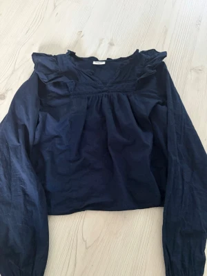 Mörkblå blus med volang från Kappahl - Snygg mörkblå blus från Kappahl med långa ärmar och volangdetaljer vid halsringningen. Blusen har en lös passform och fina spetsdetaljer framtill. Perfekt för dig som gillar en stilren och trendig look.