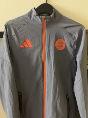 Grå  Bayern München vinddress adidas tiro - Snyggt grått träningsset från Adidas x FC Bayern München med orange detaljer och klubbmärke. Setet består av jacka med dragkedja och två fickor samt matchande byxor med dragkedjor vid fickorna. Perfekt för fotbollsträning eller chill. Materialet är lätt och funktionellt.