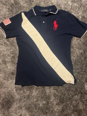 Marinblå pikétröja från Polo Ralph Lauren - Snygg marinblå pikétröja från Polo Ralph Lauren med vit diagonal rand, röd logga på bröstet och amerikansk flagga på ärmen. Klassisk krage och knappar framtill. Perfekt för en sportig och stilren look.