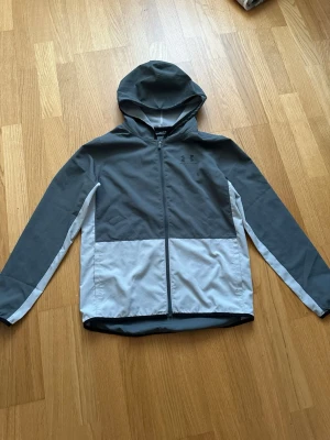 Grå och vit vindjacka från Under Armour - Snygg vindjacka från Under Armour i tvåfärgad design med mörkgrå upptill och ljusgrå nedtill. Jackan har huva, hel dragkedja framtill och smala vita detaljer längs ärmarna. Perfekt för blåsiga dagar och sportiga outfits.