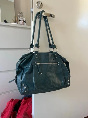 Mörkgrön handväska med silverdetaljer - Snygg mörkgrön Accessorize handväska i skinnimitation med dubbla axelband och silverfärgade metalldetaljer. Väskan har en framficka med dragkedja och dekorativa nitar i hörnen. Rymmer bra min dator som är 33cm bred så väskan passar bra till tex skolan och vardagen🥰 fråga om du har frågor!