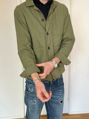 Olivgrön overshirt med fickor - Snygg olivgrön overshirt med tre stora utanpåfickor och knäppning framtill. Overshirten har klassisk krage och är tillverkad i ett kraftigare bomullstyg. Perfekt att slänga över en hoodie för en avslappnad look.