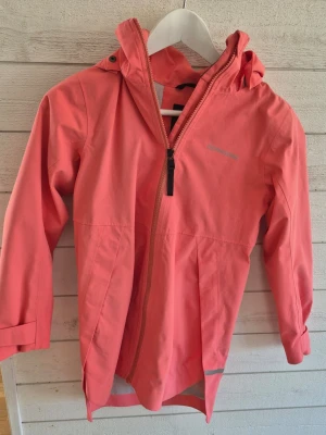 Korallrosa regnjacka från Didriksons - Snygg korallrosa regnjacka från Didriksons med huva och dragkedja framtill. Jackan är helt vattentät och har en längre modell baktill samt reflexdetalj vid nederkanten. Perfekt för regniga dagar och outdoor-äventyr.