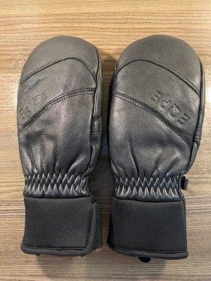 Svarta skinnvantar - EQPE - Oanvända svarta tumvantar från EQPE i skinn med breda muddar och kardborreknäppning. Snygg, stilren design med diskret logga på ovansidan, perfekt för skidbacken eller vanlig utedag