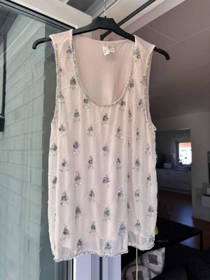 Beige paljettprydd ärmlös linnetopp/ festtopp sommartopp - Supersnygg beige ärmlös linnetopp med silverfärgade paljettdetaljer och pärlor i ett elegant mönster. Tunt, lätt material som ger en lyxig känsla och passar perfekt till ett par jeans eller kjol. Djup rund halsringning och transparent look.🥰