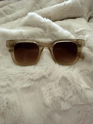Beige fyrkantiga solglasögon Chimi - Snygga solglasögon från Chimi med fyrkantig form och transparent beige båge. Glasen är mörka och bågarna har en stilren design med märkets logga på sidan. Perfekta för att lyfta din look under soliga dagar.  Det är färgen Ecru. Dom är i superfint skick