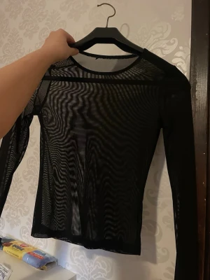 Svart mesh långärmad topp - Säljer en svart mesh topp med lång ärm och rund hals. Materialet är tunt och transparent, vilket ger en edgy och modern look. Perfekt att styla lager på lager eller som statement piece. Toppen har en normal passning och är riktigt snygg till jeans eller kjol.