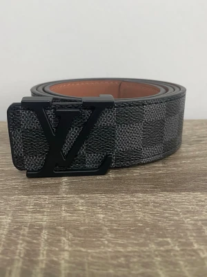 Bälte - Snyggt svart bälte med rutigt mönster och stor logga i metall på spännet. Bältet har en lyxig känsla och insidan är brun. Perfekt accessoar för att lyfta din outfit. Den har lite märken på insidan men ser helt ny ut på utsidan. Redo att skickas direkt. 