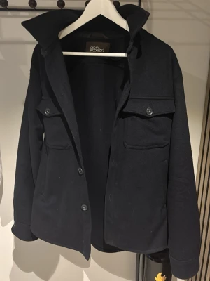 Svart overshirt från Oscar Jacobson - Stilren svart overshirt från Oscar Jacobson med två stora bröstfickor med lock och knapp, klassisk krage och knappstängning framtill. Tillverkad i ett tjockare tyg som ger en schysst och avslappnad look. Perfekt lager-på-lager-plagg.