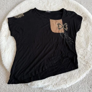 Svart topp med leoparddetaljer från Rodin - Svart topp från Rodin med korta ärmar och coola leopardmönstrade detaljer. Framtill finns en beige patch med leopardrosett och små pärlor som dekoration. Skön och avslappnad passform, perfekt till jeans eller kjol.