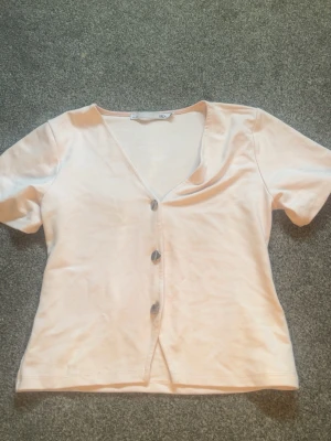 Ljusrosa topp med knappar från 157 - Söt ljusrosa topp från 157 med v-ringning och tre knappar framtill. Toppen har korta ärmar och är gjord i mjukt bomullstyg, perfekt för en avslappnad och trendig look.