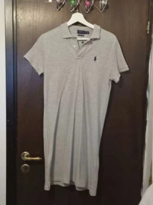 Grå pikéklänning från Ralph Lauren - Sportig grå pikéklänning från Ralph Lauren med klassisk krage, korta ärmar och knappar framtill. Klänningen har den ikoniska broderade loggan på bröstet och är gjord i mjuk bomull för en avslappnad men stilren look.