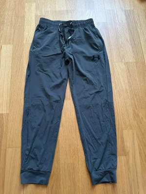 Mörkgrå mjukisbyxor från Under Armour - Snygga mörkgrå mjukisbyxor från Under Armour med resår och dragsko i midjan. Byxorna har smal passform, muddar vid bensluten och två sidofickor. Perfekta för chill eller träning. Materialet är mjukt och lätt för extra komfort.