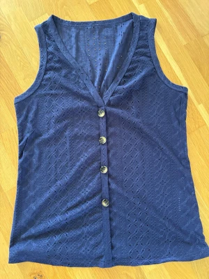 Linne med spets mönster  - Ärmlöst linne med V-ringning och dekorativa knappar framtill. Tyget har ett fint hålmönster/spetsliknande struktur som ger ett somrigt och lite elegant uttryck. Perfekt till vardag och finare tillfällen. 