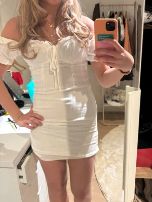 Vit off shoulder miniklänning - Säljer en vit, tight off shoulder miniklänning med korta puffärmar och snörning framtill. Klänningen har en figurnära passform och lätt ribbad struktur. Perfekt för dig som gillar en trendig och feminin stil. Perfekt till skolavslutning eller student! Använd 1 gång