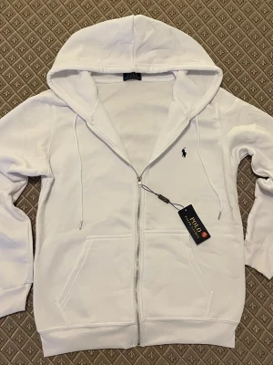 Vit hoodie från Polo Ralph Lauren - Klassisk vit hoodie från Polo Ralph Lauren med dragkedja framtill, huva med snörning och två stora fickor. Ikoniska loggan broderad på bröstet i svart. Tillverkad i mjuk bomull för en skön och avslappnad look.